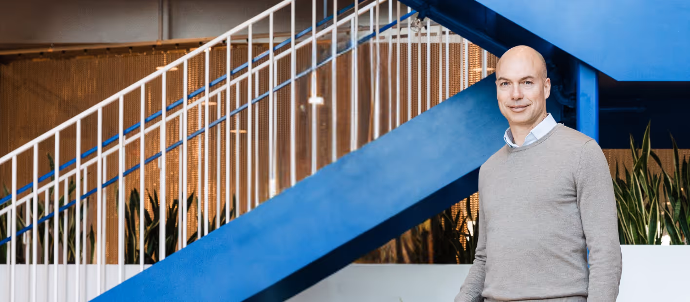 Bald man wearing a light gray sweater standing indoors near a staircase with blue and white railings and plants in the background.