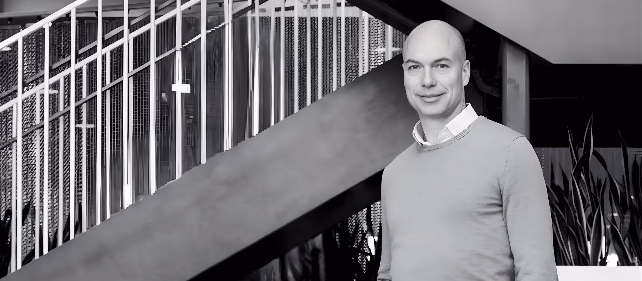 Bald man wearing a light gray sweater standing indoors near a staircase with blue and white railings and plants in the background.