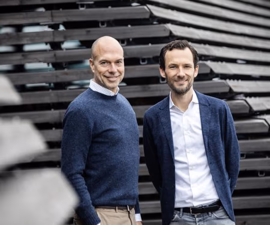 Two men standing and smiling in front of a modern architectural wall with horizontal black panels.