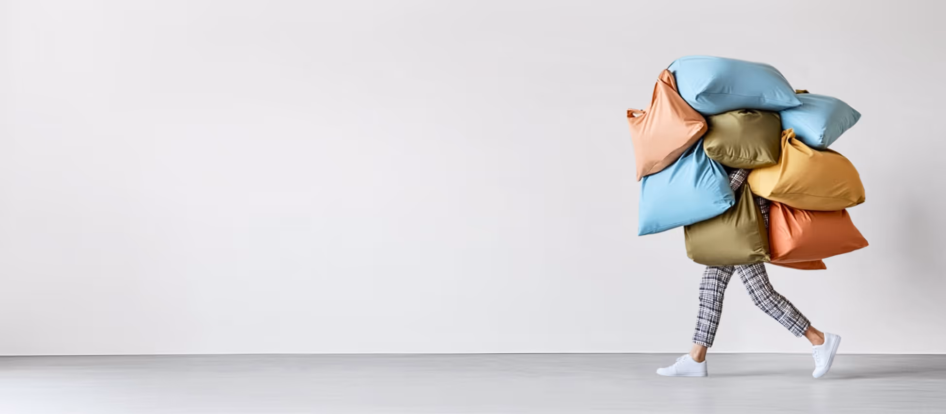 Person walking carrying a large stack of colorful pillows, wearing plaid pants and white sneakers.