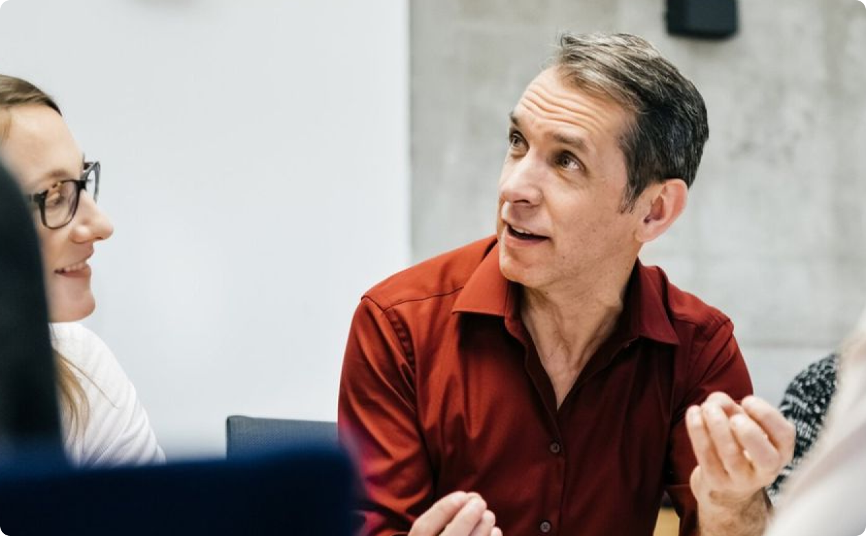 Man in red shirt engaged in conversation with woman wearing glasses in an office setting.