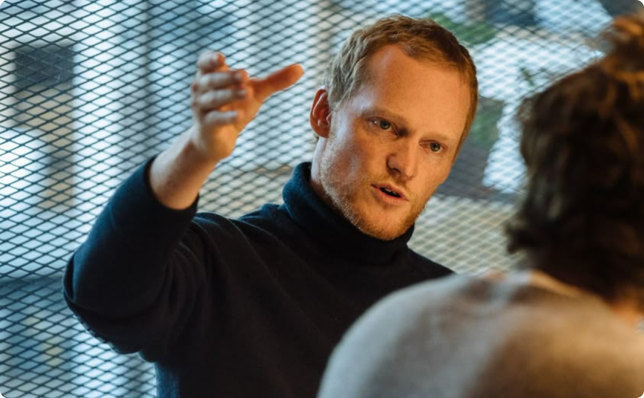 Man in a black turtleneck speaking with expressive hand gesture to a woman with curly hair in front of a mesh screen.