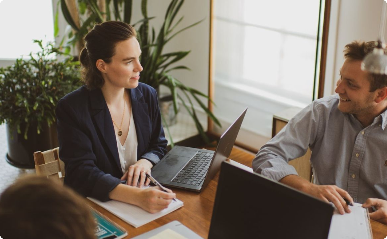 A woman and a man sitting at a wooden table with laptops and notebooks, engaged in a discussion in a bright room with plants.