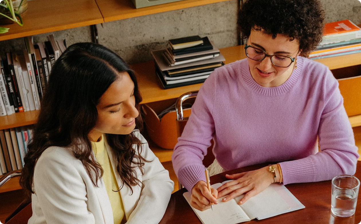 Two women sitting at a table with one writing in a notebook and the other looking on attentively in a cozy room with bookshelves.