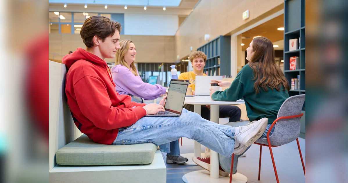 Students in casual attire study and chat in a modern library, with one on a laptop. Bright, airy atmosphere with bookshelves visible.