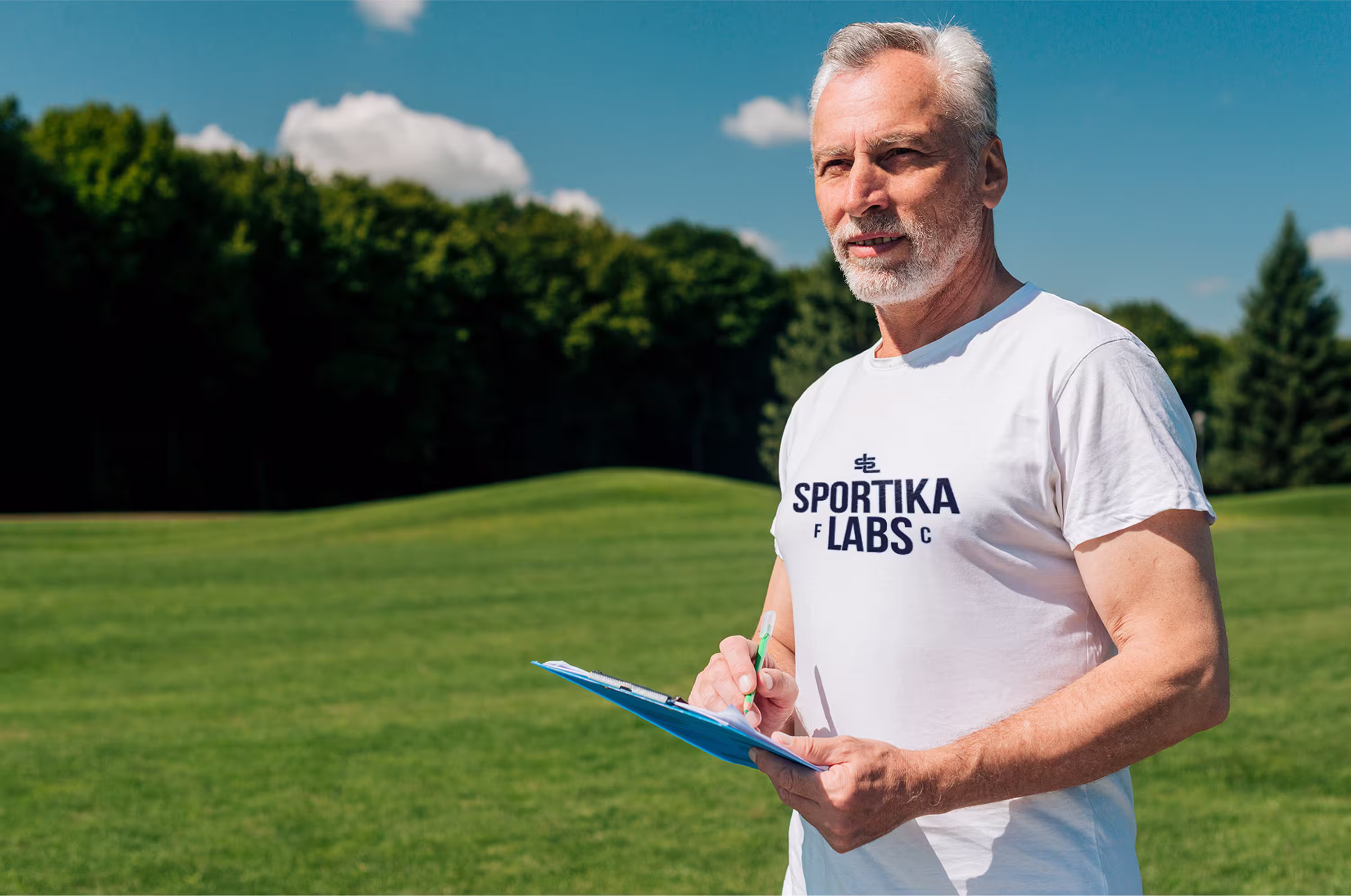 Senior coach holding a clipboard and standing on the soccer field under a blue sky, wearing a Sportika Labs T-shirt.