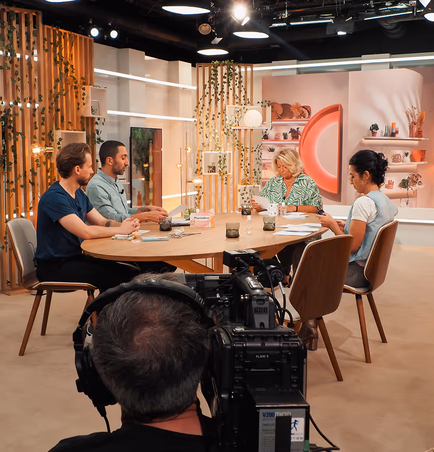 A television studio set featuring four people sitting around a circular table equipped with coffee cups and notebooks. The backdrop has decorative shelves and plants, with a camera in the foreground focusing on the participants.