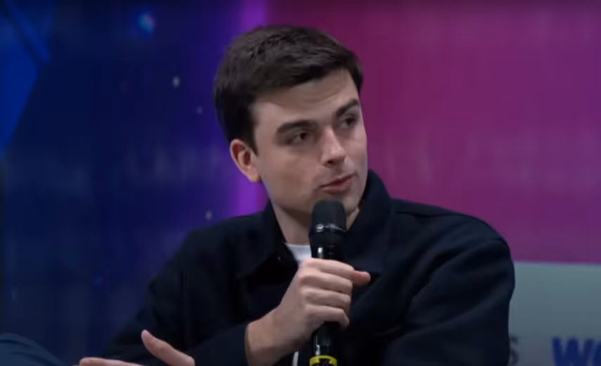 A person speaking into a microphone during a panel discussion, wearing a black jacket and white shirt, with a colorful background.