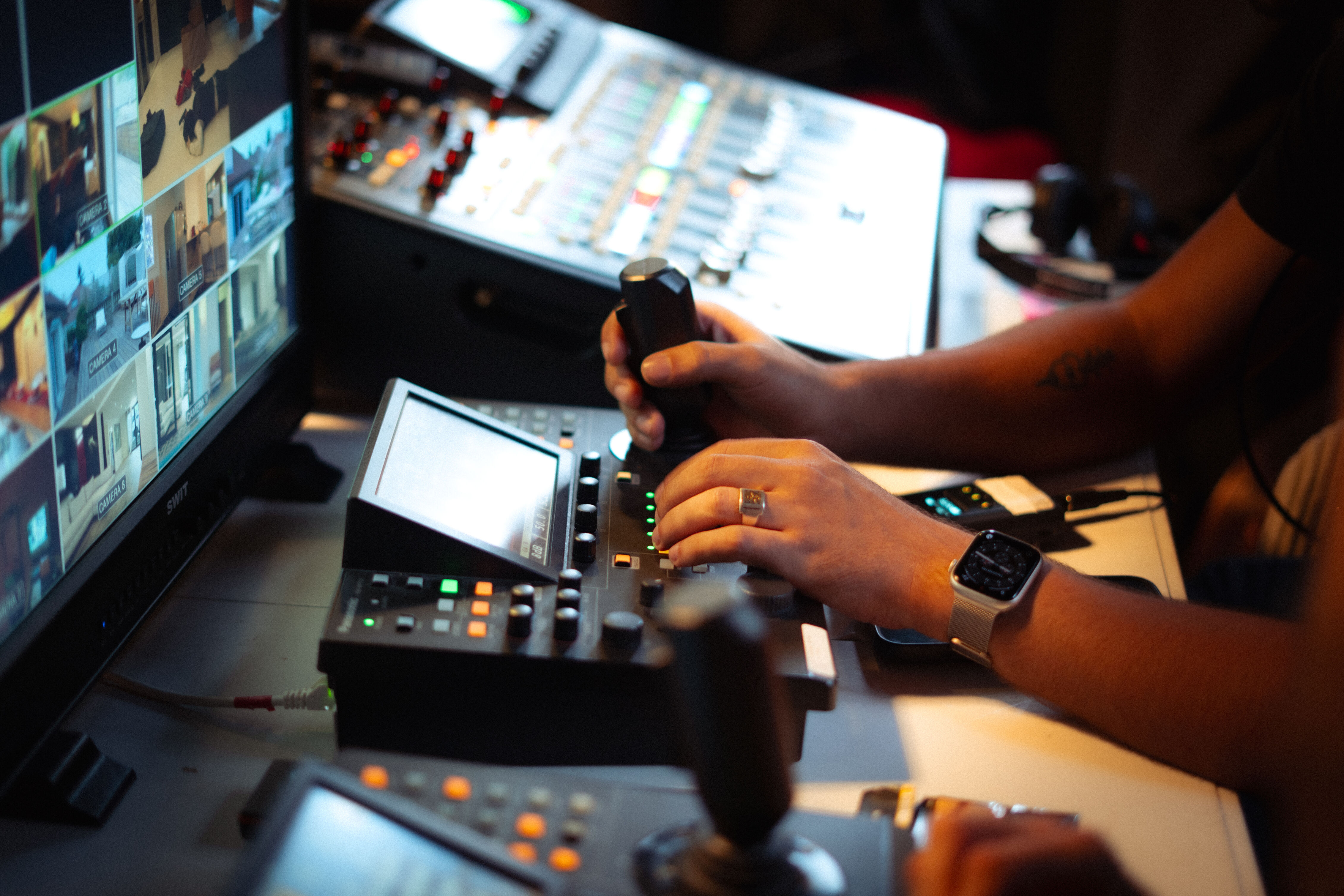 Two men working with video production equipment in a control room. One man is operating a control panel while looking at multiple monitors displaying a news presenter