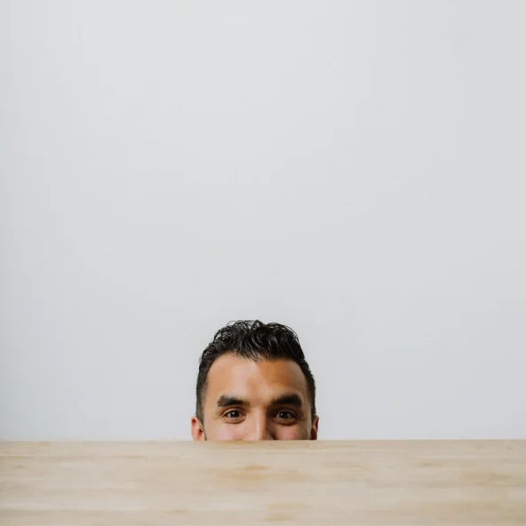 Man peeks playfully over the edge of a wooden table, with only his eyes and forehead visible against a plain wall.