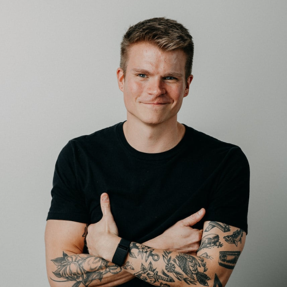 Smiling man with short hair and tattooed arms crosses his arms while wearing a black T-shirt.