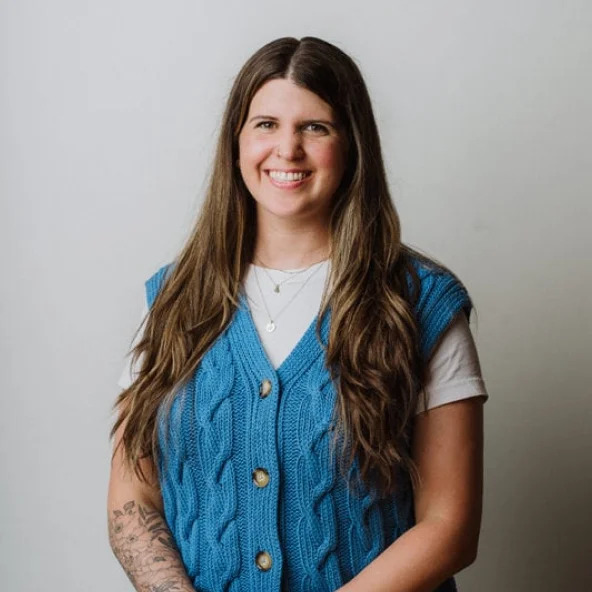 Smiling woman in a blue cable-knit vest and white T-shirt sits at a wooden table with tattooed arm visible.