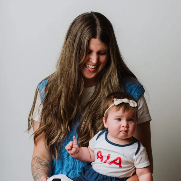 Smiling mother holds baby wearing a white outfit with a bow and soccer ball on a wooden table.