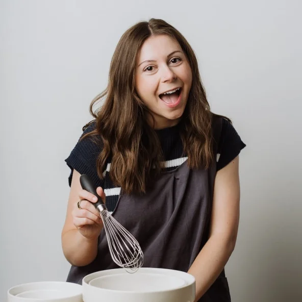 Smiling woman holding a whisk over mixing bowls on a wooden table with cookies and baking tools.