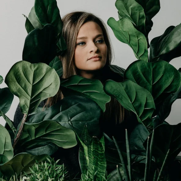 Young woman surrounded by lush green indoor plants looks thoughtfully to the side.