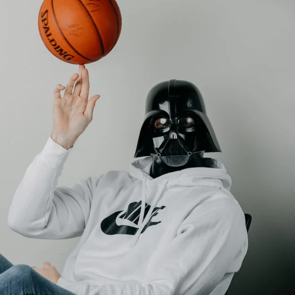 Person wearing a Darth Vader mask and white Nike hoodie spins a basketball on one finger while sitting at a table.