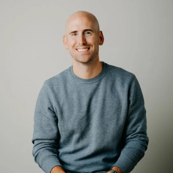 Smiling bald man in a gray sweatshirt sits at a wooden table with hands clasped.