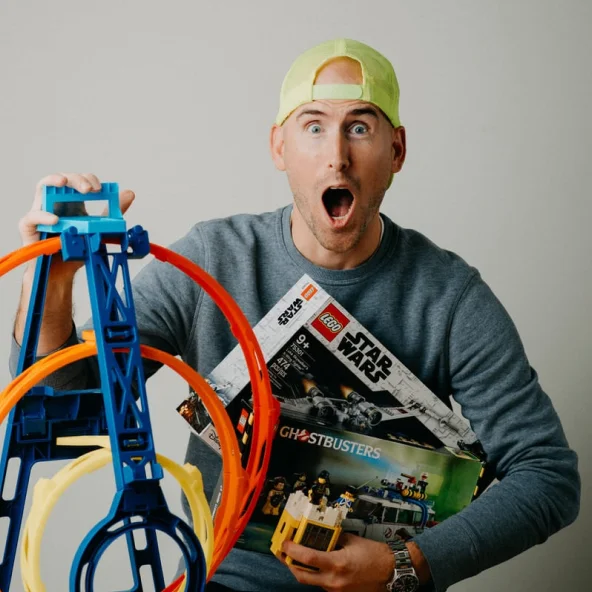 Excited man wearing a backwards neon cap holds LEGO Star Wars and Ghostbusters sets beside a Hot Wheels track.