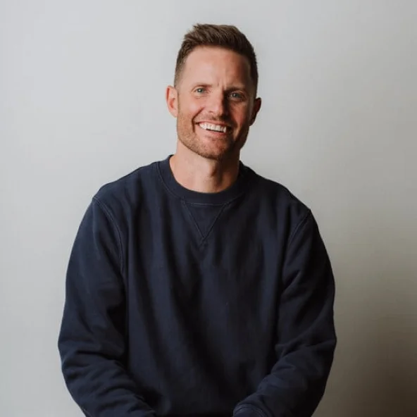 Man in a navy sweatshirt smiles while sitting at a table with hands resting in front.