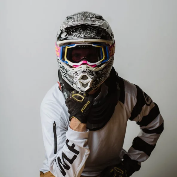 Motocross rider in helmet and goggles rests chin on gloved hand while seated at a wooden table.
