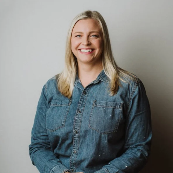 Smiling woman in a denim shirt sits at a wooden table with hands folded.