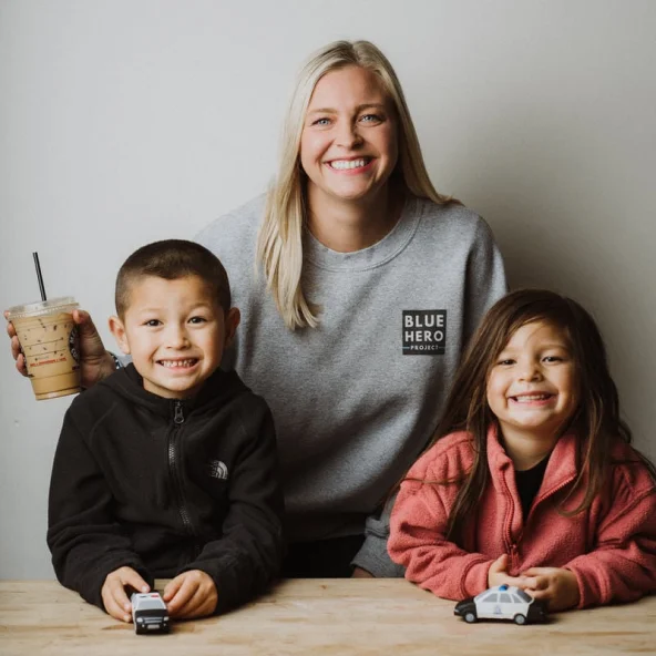 Woman with two smiling children sits at a table holding iced coffee while the kids play with toy cars.