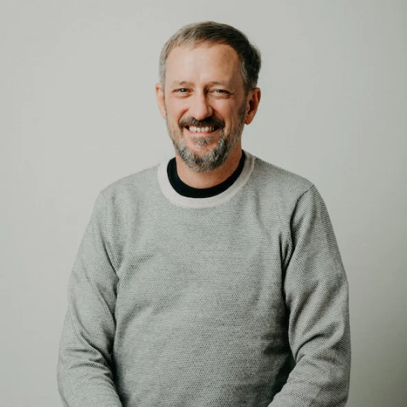 Man with short gray hair and beard smiling while wearing a light gray sweater, seated at a wooden table.