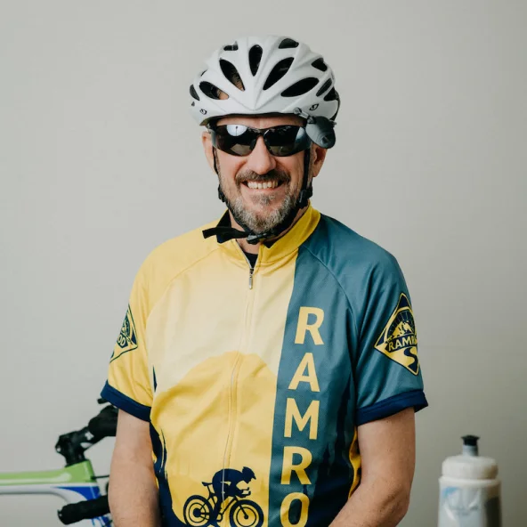 Man wearing a yellow and blue cycling jersey, helmet, and sunglasses sits beside a bike with a water bottle on the table.