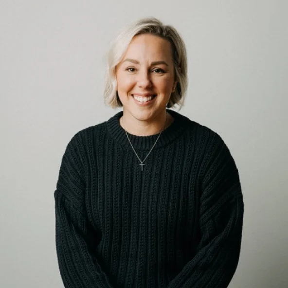 Smiling woman with short blonde hair wearing a black ribbed sweater and a cross necklace, seated at a wooden table.