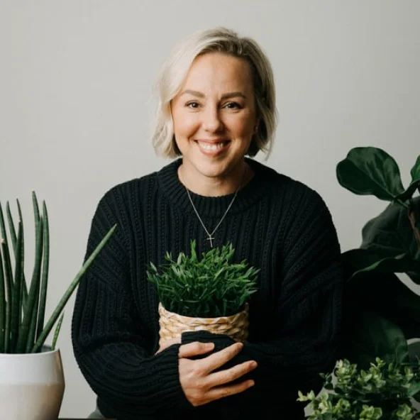 Smiling woman in a black sweater holds a potted plant at a wooden table surrounded by other plants and books.