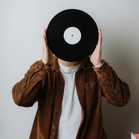 Person wearing a brown corduroy shirt holds a vinyl record in front of their face, surrounded by stacks of albums on a table.
