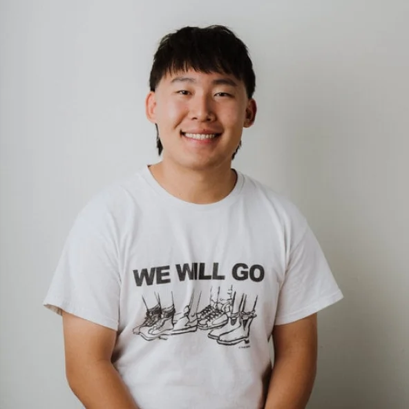 Smiling young man wearing a white “We Will Go” T-shirt sits at a wooden table.