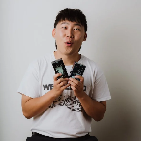 Man in a white T-shirt holds two guitar pedals with several other effects pedals arranged on the table.