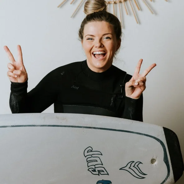 Smiling woman in a wetsuit flashes peace signs while holding a surfboard in front of a decorative wall mirror.