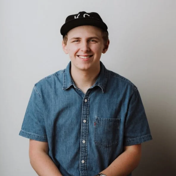Smiling young man in a denim shirt and black cap sits at a wooden table with hands clasped.