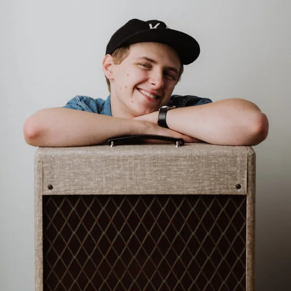 Smiling young man wearing a black cap leans on a guitar amplifier placed on a wooden table.