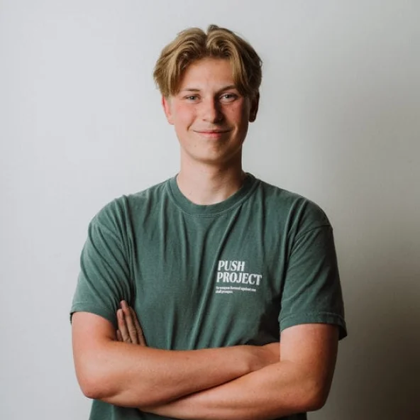 Young man wearing a green Push Project T-shirt smiles with arms crossed at a wooden table.