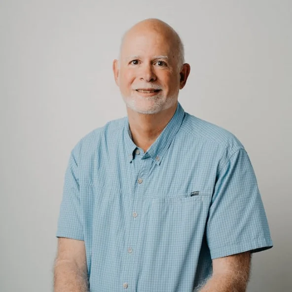 Older man with a trimmed beard and light blue short-sleeve shirt smiles while sitting at a wooden table.