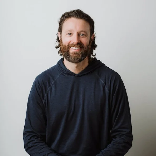 Smiling man with medium-length hair and beard in a navy hoodie sits at a wooden table.