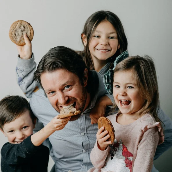 Man playfully bites a cookie while laughing with three children, all holding large cookies together.