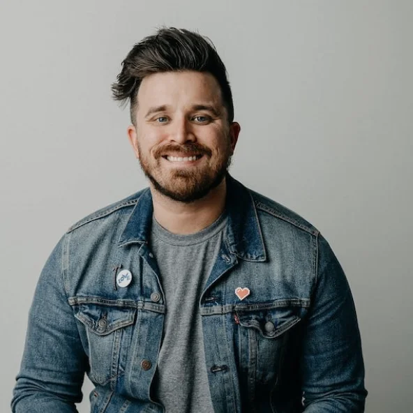 Smiling man with styled hair and beard wearing a denim jacket and gray shirt sits at a wooden table.