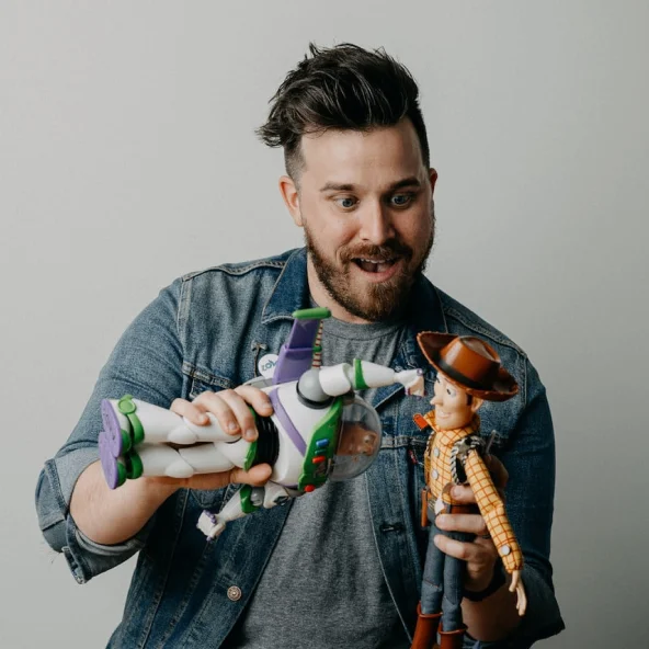 Man with excited expression holds Buzz Lightyear and Woody action figures surrounded by collectible figurines on a table.