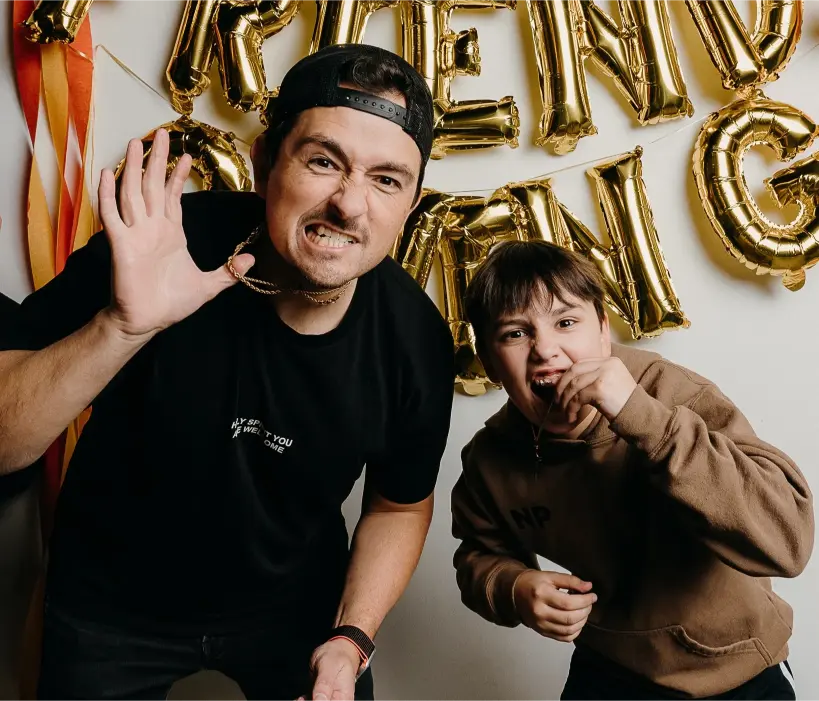 Man and boy posing playfully in front of gold letter balloons, both smiling with fun expressions.
