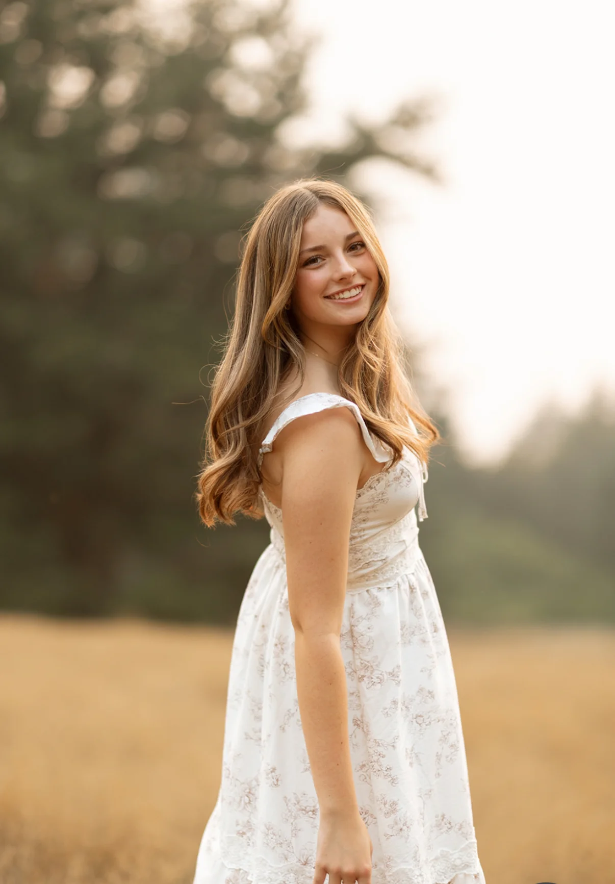 Smiling woman with long light brown hair wearing a white patterned dress in a sunny outdoor field.