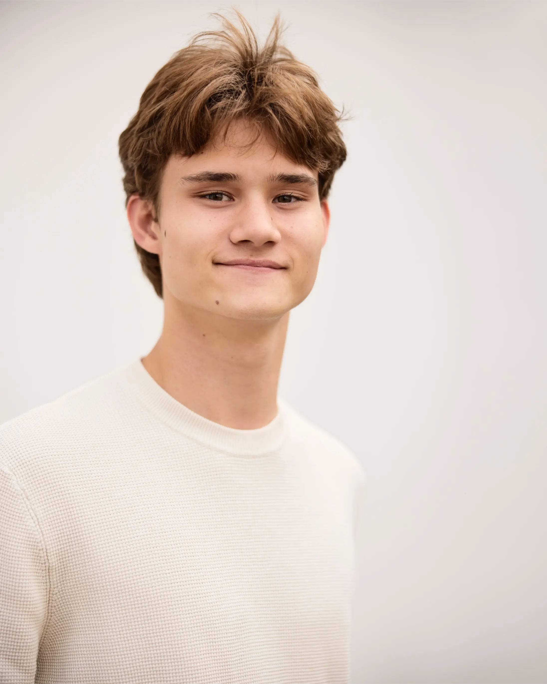 Young man with brown hair wearing a cream-colored sweater, smiling slightly against a light background.