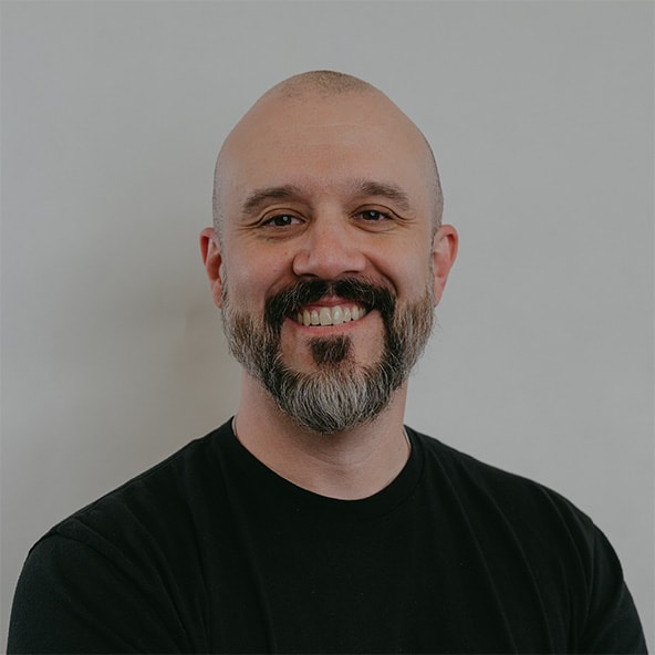 Smiling man with beard wearing a black shirt against a grey background.