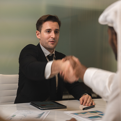 Two businessmen shaking hands across a table with documents and a tablet during a meeting.