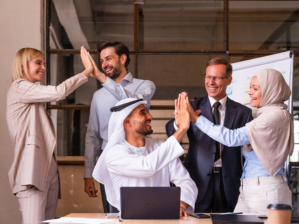 Diverse group of five business professionals smiling and giving high fives in an office setting.