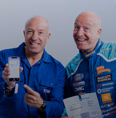 Two smiling bald men, one in a blue shirt holding a smartphone and the other in a racing suit holding documents.