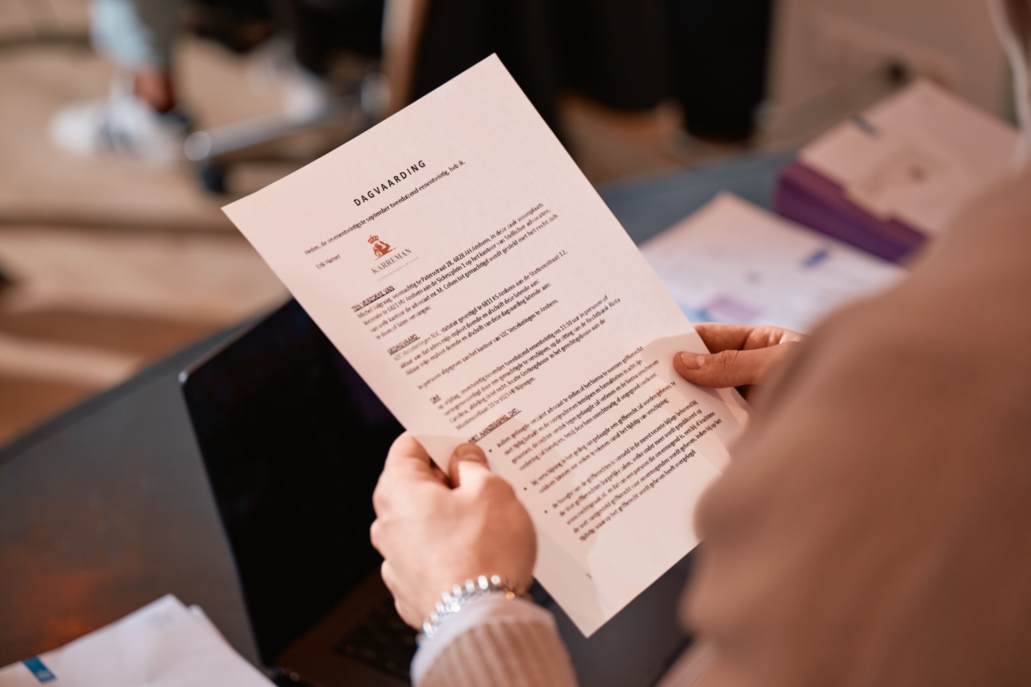 Person holding and reading a printed sheet of paper with text titled 'DAGVAARDING' over a laptop on a desk.